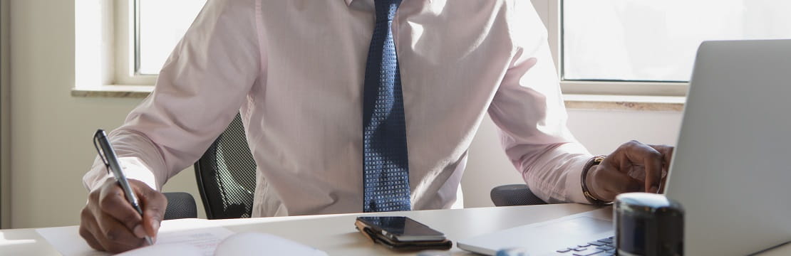 Businessman sits at a desk facing a computer and writing on a sheet of paper